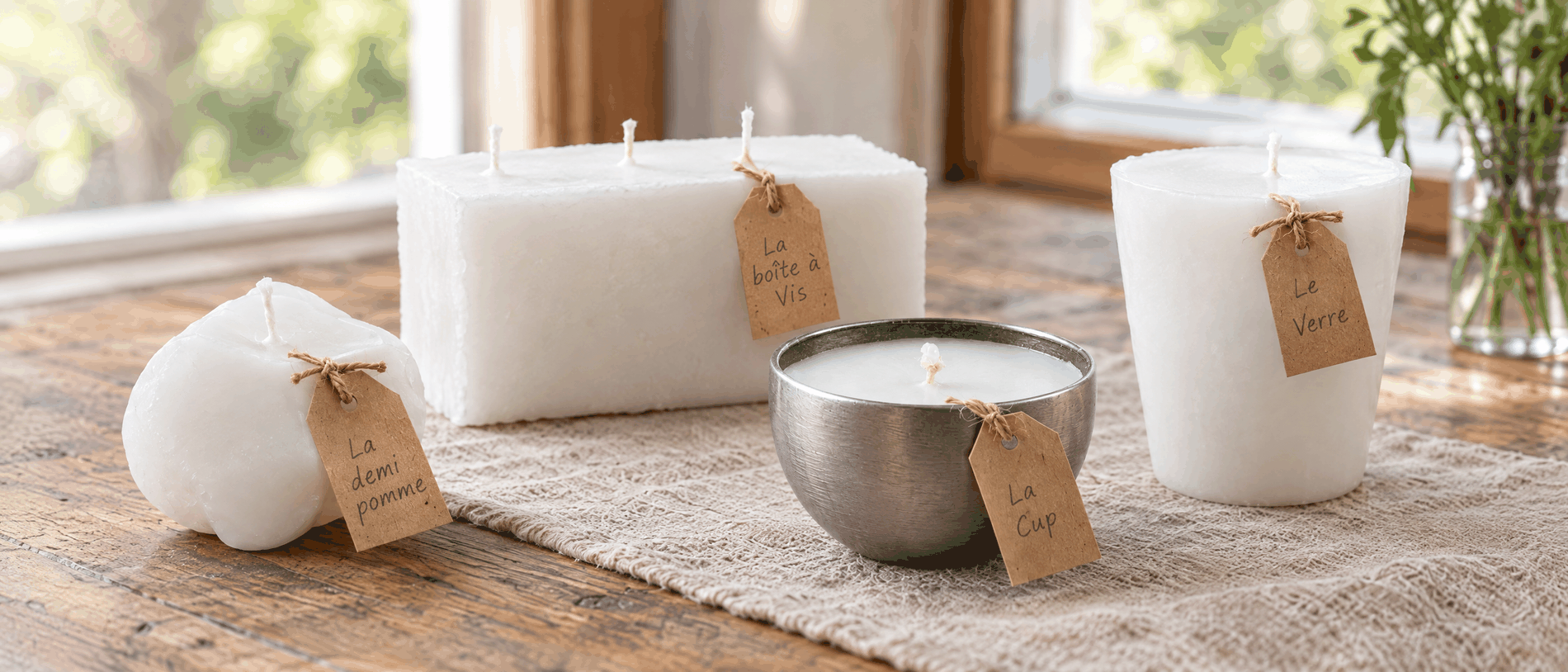 Quatre chandelles blanches posées sur une table de bois, chacune avec une étiquette kraft écrite à la main&nbsp;: La demi pomme, La boîte à Vis, La Cup et Le Verre